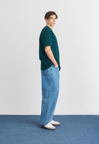 Πράσινο βαμβακερό t-shirt με άνετη εφαρμογή, σε συνδυασμό με ανοιχτό μπλε τζιν παντελόνι με φαρδύ πόδι. Το μοντέλο φοράει λευκά αθλητικά παπούτσια και στέκεται σε ένα μπλε χαλί.