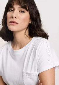 Biała koszulka z miękkiej bawełny, z kieszonką i ozdobnymi ćwiekami na ramionach. Prosty okrągły dekolt i krótkie rękawy.