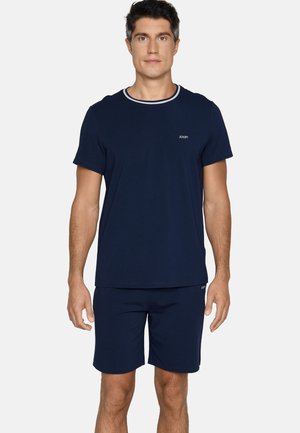 Mann trägt ein dunkelblaues Kurzarmhemd mit weißem Kragenbesatz und passende Shorts, steht vor einem schlichten Hintergrund.
