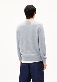 Grauer Sweatshirt mit gerippten Bündchen und Saum, strukturiertem Stoff und einem kleinen Etikett am Kragen, getragen über dunkelblauen Hosen.