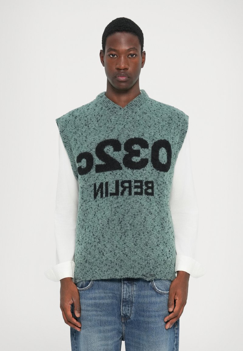 Maglione senza maniche lavorato a maglia in teal con una finitura testurizzata. Presenta un testo nero in grassetto "032 BERLIN" sulla parte anteriore. Indossato sopra una camicia bianca a maniche lunghe.