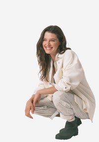 Donna sorridente con lunghi capelli castani in posizione accovacciata, che indossa una giacca crema, pantaloni chiari a fantasia e stivali neri alla caviglia su uno sfondo neutro.