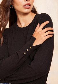 Pulover negru cu decolteu rotund și mâneci lungi. Are trei nasturi decorativi aurii pe fiecare manșetă. Material moale, texturat.