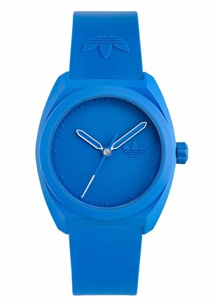Blue silicone watch with a round face, textured dial, minimalist hour markers, and white hands. Features a button on the side and logo on the strap.
