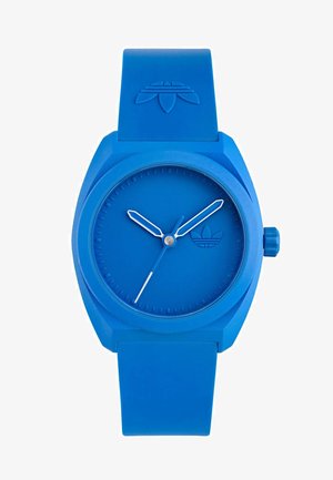 Modra silikonska ura z okroglo obliko, teksturiranim številčnico, minimalističnimi označevalci ur in belimi kazalci. Ima gumb na strani in logo na traku.