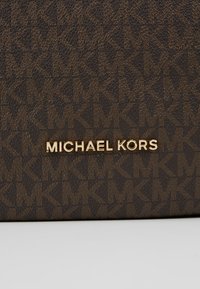 Surface texturée marron avec un motif répété "MK" et un lettrage doré "MICHAEL KORS" centré sur le tissu.