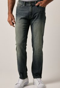 Jeans de mezclilla azul oscuro con un lavado desgastado, diseño de cinco bolsillos y costuras en contraste. Combinados con zapatillas blancas.
