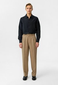 Zwarte overhemd met knopen en een gestructureerde afwerking, gecombineerd met beige hoge taille broek. Zwarte schoenen maken de look compleet. Effen achtergrond.