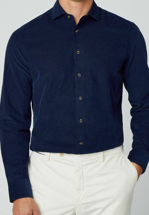 Hombre con camisa azul marino de manga larga abotonada, metida dentro de pantalones blancos a medida, con las manos en los bolsillos.