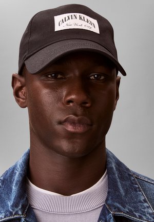 Noor mees, kandes musta Calvin Kleini nokamütsi, valget soonikut särki ja sinist teksajopet, seisab halli tausta ees neutraalse ilmega otse vaataja poole.