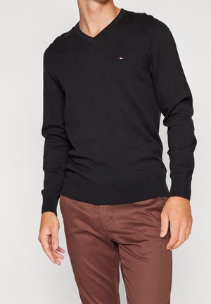 Mężczyzna w czarnym swetrze z dekoltem w serek i małym logo na piersi oraz brązowych spodniach, stojący na tle jednolitego jasnego tła.