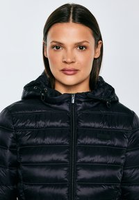 Vrouw met steil donker haar, gekleed in een zwarte gewatteerde jas met ritssluiting en capuchon, kijkt recht vooruit tegen een effen lichte achtergrond.