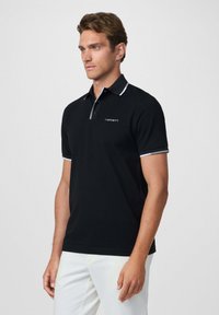 Camisa polo negra con un cuello y puños de contraste, con sutiles detalles en gris y un logo bordado en el pecho. Corte ajustado.