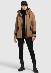 Bruine gevoerde jas met een capuchon, zwarte accenten en trekkoorden. Draag deze over een zwarte coltrui en zwarte leggings met tekst, gecombineerd met zwarte laarzen.