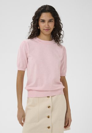 Jeune femme aux cheveux bouclés bruns portant un pull rose à manches courtes et une jupe beige boutonnée sur le devant, debout devant un fond gris.