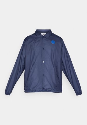 Giacca a vento blu navy realizzata in materiale leggero. Presenta una chiusura a bottoni, un'etichetta con logo blu e polsini elastici.