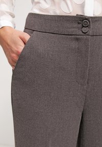 Pantalon gris sur mesure avec une texture lisse, doté d'une fermeture à boutons et de poches latérales, associé à un chemisier transparent orné de motifs circulaires.