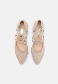 Chaussures à talons aiguilles en suede beige avec un bout pointu, comportant deux brides ajustables croisées sur le cou-de-pied et un logo métallique poli sur la semelle intérieure.