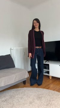 Egy nő áll egy nappaliban fekete ingben, bordó kardigánban, bőszárú farmerral és barna övvel, miközben mosolyog a kamerába.