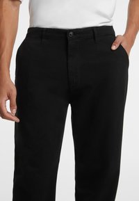Schwarze Hose mit geradem Bein, ausgestattet mit einem Knopfverschluss, Gürtelschlaufen und seitlichen Taschen. Hergestellt aus glattem Stoff mit einem sauberen Finish.