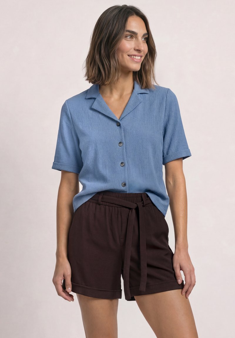Femme souriante, portant une chemise bleue à manches courtes boutonnée et un short marron foncé avec un lien à la taille, debout devant un fond uni.