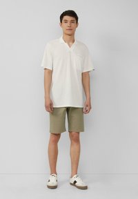 Weißes, strukturiertes Kurzarm-Polohemd mit einer Brusttasche, kombiniert mit olivgrünen Shorts und weißen Sneakers mit dunklen Akzenten.