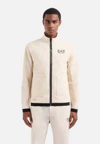 Giacca beige con zip, dotata di due tasche frontali, dettagli neri e un logo sul petto. Materiale liscio con una vestibilità strutturata.