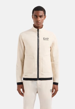 Giacca beige con zip, dotata di due tasche frontali, dettagli neri e un logo sul petto. Materiale liscio con una vestibilità strutturata.