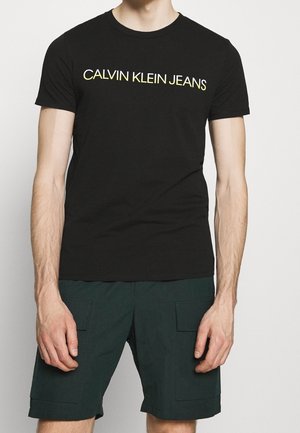 Mand iført sort Calvin Klein Jeans t-shirt og mørkegrønne cargo shorts, stående med armene afslappet ned langs siden.
