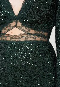 Πράσινο φόρεμα με παγιέτες που έχει βαθύ V-ντεκολτέ και δαντελένιες λεπτομέρειες στη μέση, με υφή που λάμπει και περίπλοκες δαντελένιες λεπτομέρειες.