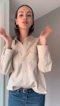 Jeune femme aux cheveux foncés porte une chemise beige boutonnée, des boucles d’oreilles créoles dorées et un jean bleu, levant les mains dans un cadre intérieur.
