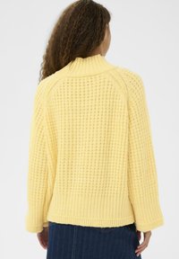 Femme aux longs cheveux bouclés portant un pull en tricot jaune texturé et un pantalon bleu foncé, vue de dos sur un fond uni.