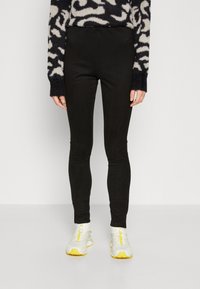 Black leggings made from stretchy material, featuring a slim fit and ankle length. Paired with light-colored athletic shoes.