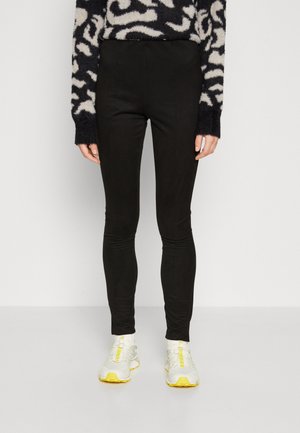 Black leggings made from stretchy material, featuring a slim fit and ankle length. Paired with light-colored athletic shoes.