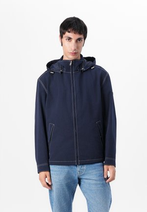 Navy blauwe zip-up jas met een capuchon, voorzien van contrasterende witte stiksels, twee zijzakken en een ontspannen pasvorm.