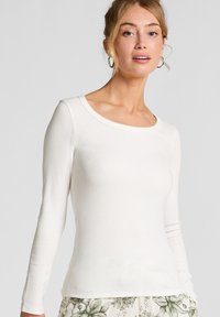 Jong meisje met een lichte huid en blond haar draagt een nauwsluitend wit T-shirt met lange mouwen en een broek met een bloemenpatroon, terwijl ze tegen een effen achtergrond staat.