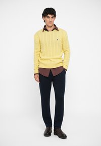 Dzeltena adīta džemperis, brūna rūtainā krekla apakšā, tumši bikses un brūnas kurpes. Modelis stāv pretī vienkāršai fona.