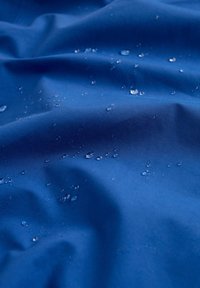 Vandeniui atsparus mėlynas audinys su lygia tekstūra, ant paviršiaus matomi vandens lašai, pabrėžiantys jo drėgmės atstūmimo savybes.