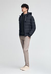 Czarna kurtka puchowa z kapturem i akcentami logo, zestawiona z teksturowanym swetrem, jasnobeżowymi spodniami i jasnymi sneakersami.