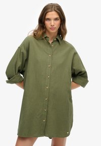 Camisa de gran tamaño en tejido verde oliva, con un frente abotonado, mangas enrolladas y un pequeño logotipo en el dobladillo. Textura suave.