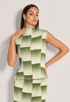 Grünes, plissiertes Oberteil mit hohem Kragen, verziert mit einem geometrischen Muster in verschiedenen Grüntönen. Akzentuiert durch große, bunte Ohrringe.