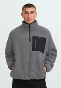 Grauer Fleece-Pullover mit hohem Kragen und einer schwarzen Brusttasche. Verfügt über einen Frontreißverschluss, lange Ärmel und einen strukturierten, gemütlichen Stoff.