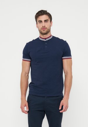 Férfi, aki testhez simuló sötétkék rövid ujjú galléros pólót visel, melynek gallérja és ujjvége csíkos piros, fehér és kék színben, fehér háttér előtt áll.