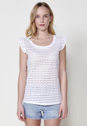 Top in cotone bianco caratterizzato da un design perforato, maniche corte a volant e scollatura a girocollo. Abbinato a pantaloni shorts in denim.