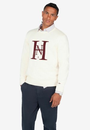 Jersey crema con el logo "HN" en color burdeos, con puños y dobladillo acanalados. Llevado sobre una camisa a rayas azul clara, combinado con pantalones azul marino oscuros.