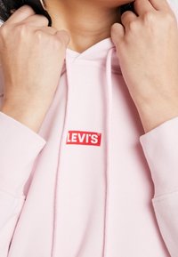 Persona viste una sudadera de Levi's rosa, sujetando los cordones de la capucha cerca de su cuello con ambas manos.