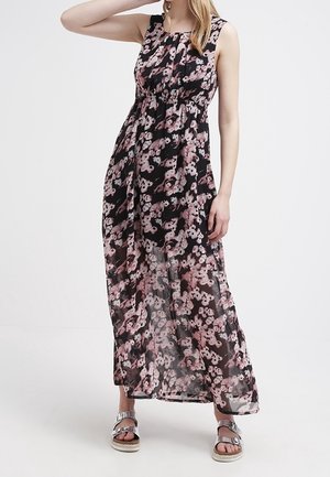 Femme portant une robe maxi noire sans manches avec un imprimé floral rose et des sandales à plateforme ouvertes argentées, se tenant devant un fond blanc.