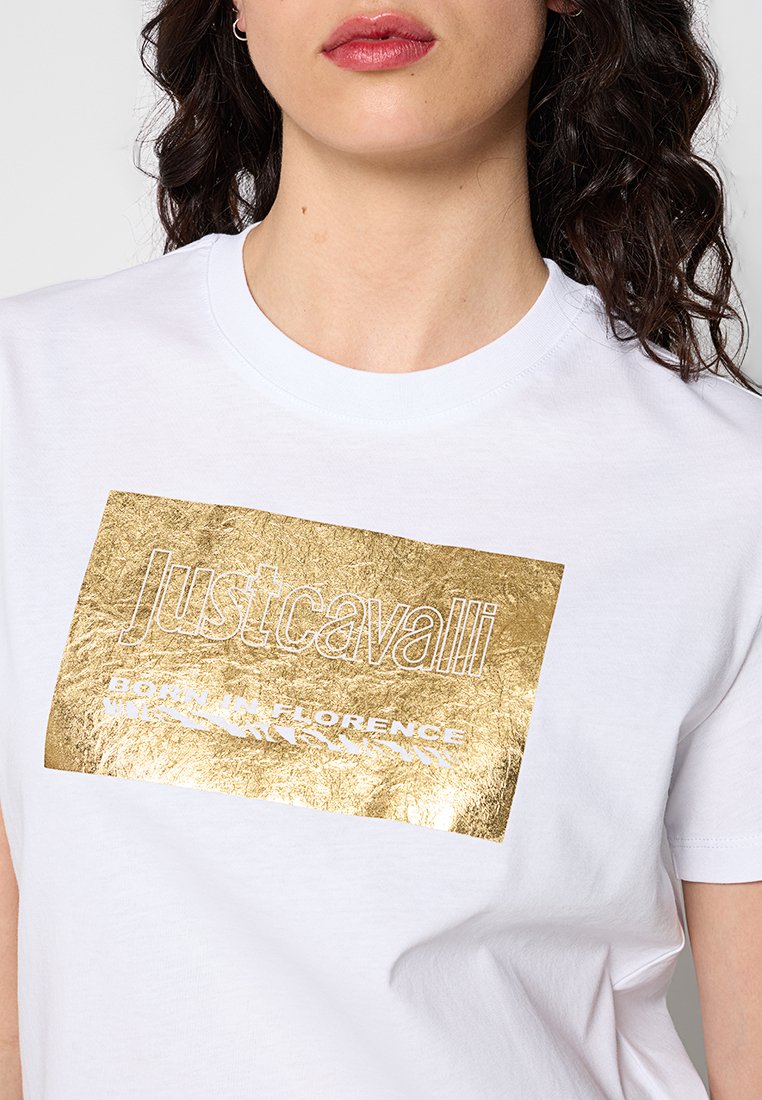 Mujer vistiendo una camiseta blanca con un logo rectangular dorado que dice "Just Cavalli" y "Born in Florence" en el pecho.