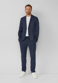 Dunkelblauer Anzug mit tailliertem Blazer und Jogginghosen mit Zugkordel, getragen über einem weißen T-Shirt. Kombiniert mit weißen Turnschuhen, die ein lässiges Design unterstreichen.