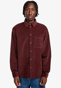 Camisa de pana burdeos con cuello abotonado, bolsillo frontal y botones marrones. Mangas largas y corte relajado. Sin patrones visibles.